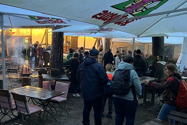 Menschen stehen im Freien unter Zelten, eine Gruppe sitzt an Tischen, im Hintergrund brennt ein Feuer.