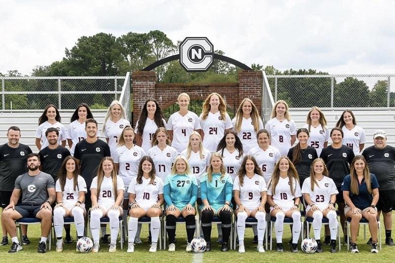 Mannschaftsfoto einer Frauenfußballmannschaft in weißen Trikots auf einem Sportplatz, begleitet von Trainern.