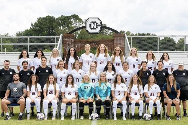 Mannschaftsfoto einer Frauenfu&szlig;ballmannschaft in wei&szlig;en Trikots auf einem Sportplatz, begleitet von Trainern.
