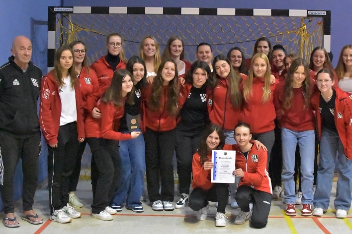 Gruppenfoto einer weiblichen Handballmannschaft in roten Trainingsanz&uuml;gen mit Trainer, Medaille und Urkunde in der Halle.