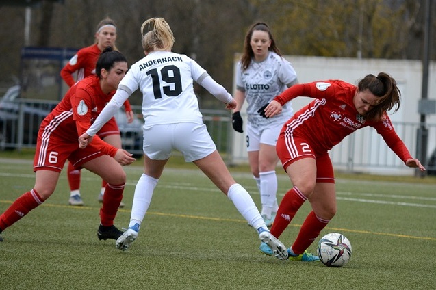Drei Fu&szlig;ballspielerinnen im Wettkampf um den Ball auf einem Rasenplatz, eine in wei&szlig;, zwei in rot.