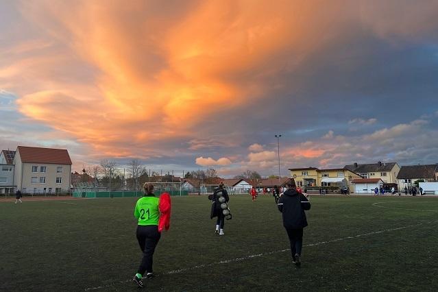 Spielerinnen verlassen den Fußballplatz bei Sonnenuntergang, beeindruckender Himmel mit orangefarbenen Wolken.