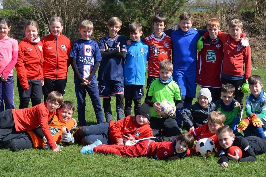Gruppe von 16 Kindern in Sportanz&uuml;gen auf einem Fu&szlig;ballfeld, einige kniend, andere liegend, mit B&auml;llen in der Mitte.