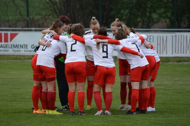Eine Fu&szlig;ballmannschaft von Frauen steht im Kreis und umarmt sich vor dem Spiel auf dem Rasenplatz.