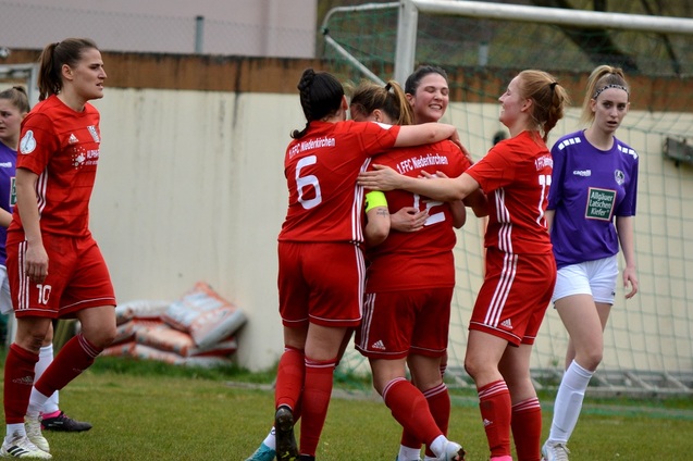 Spielerinnen in roten Trikots feiern einen Torerfolg auf dem Fu&szlig;ballfeld, w&auml;hrend eine Spielerin in lila zusieht.