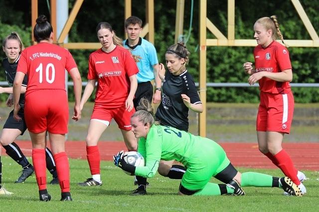 Eine Torwartspielerin in gr&uuml;ner Ausr&uuml;stung f&auml;ngt den Ball, w&auml;hrend Spielerinnen in roten Trikots angreifen.