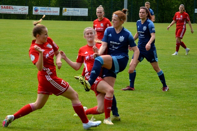 Zwei Fu&szlig;ballspielerinnen im roten Trikot k&auml;mpfen um den Ball, w&auml;hrend weitere Spielerinnen im Hintergrund zuschauen.