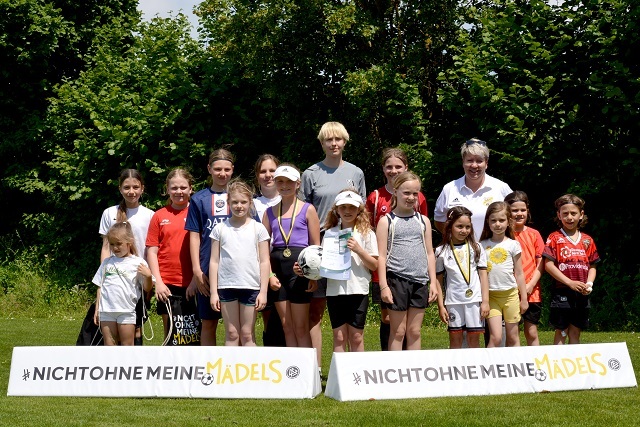 Gruppe von M&auml;dchen und Frauen steht auf einem Sportplatz vor Bannern mit dem Slogan &bdquo;Nicht ohne meine M&auml;dels&ldquo;.