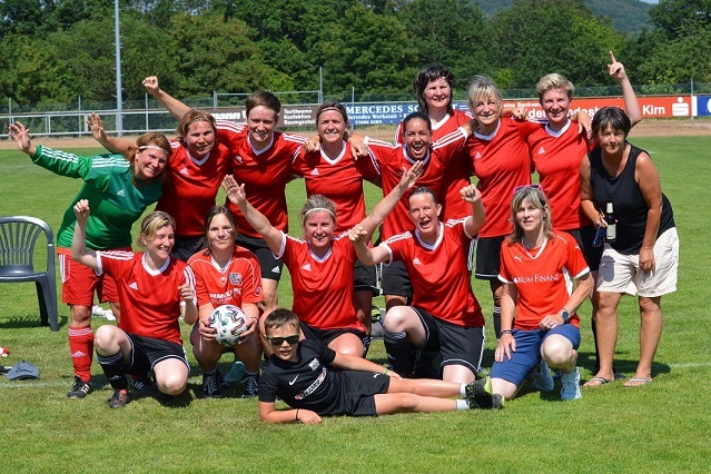 Gruppenshot eines Frauenfu&szlig;ballteams in roten Trikots, freudig posierend auf einem Sportplatz mit gr&uuml;nem Hintergrund.