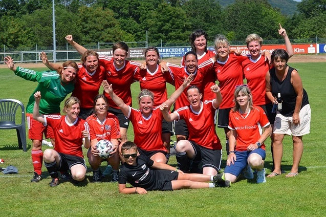 Gruppenshot eines Frauenfu&szlig;ballteams in roten Trikots, freudig posierend auf einem Sportplatz mit gr&uuml;nem Hintergrund.