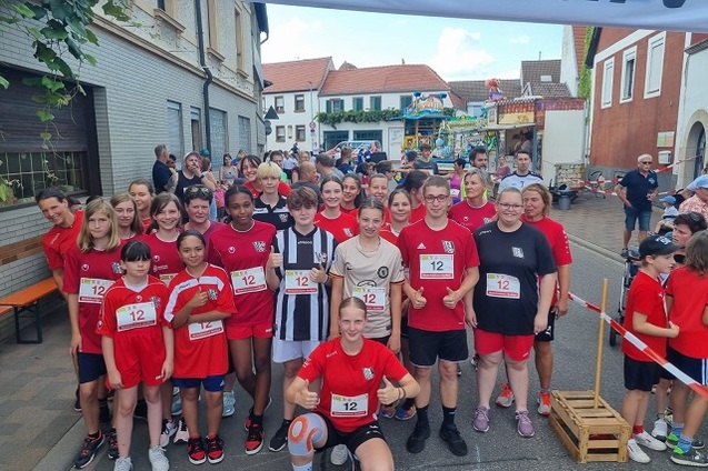 Gruppe junger Sportler in roten-Shirts stehen unter einem Startbanner, bereit f&uuml;r ein Rennen in einer Stadtstra&szlig;e.