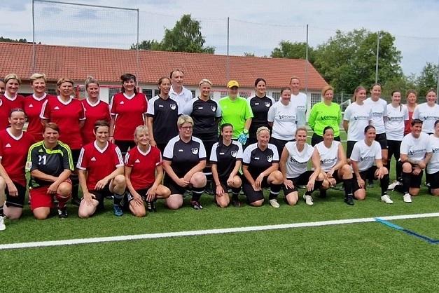 Gruppenfoto von 25 Frauen in Fußballtrikots, auf einem Sportplatz mit Rasen und Toren im Hintergrund.