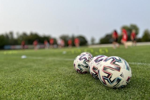Nahaufnahme von drei Fußbällen auf einem grünen Rasenfeld mit Spielern im Hintergrund, die trainieren.