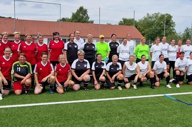 Gruppenfoto von 25 Frauen in Fu&szlig;balltrikots, auf einem Sportplatz mit Rasen und Toren im Hintergrund.