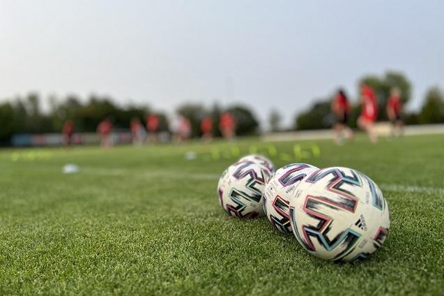 Nahaufnahme von drei Fu&szlig;b&auml;llen auf einem gr&uuml;nen Rasenfeld mit Spielern im Hintergrund, die trainieren.