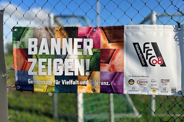 Banner mit dem Text "Banner zeigen! Gemeinsam f&uuml;r Vielfalt und Toleranz." an einem Zaun. Farbige Hintergr&uuml;nde in Sektionen.