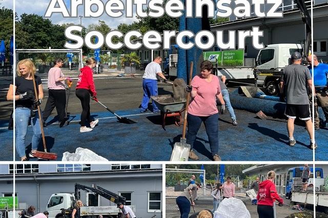 Volunteers arbeiten am Soccercourt, schieben Material und nutzen Werkzeuge unter blauem Himmel.