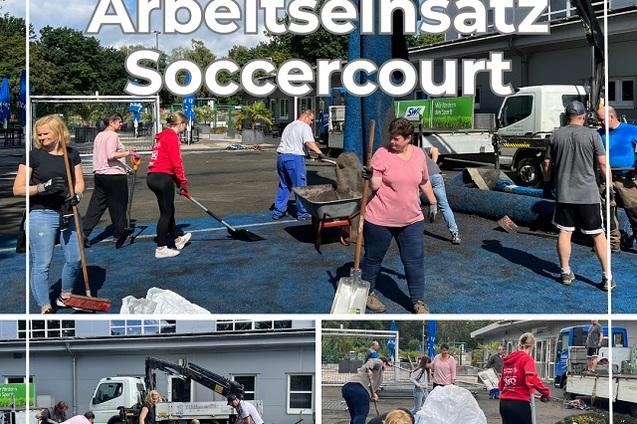 Volunteers arbeiten am Soccercourt, schieben Material und nutzen Werkzeuge unter blauem Himmel.