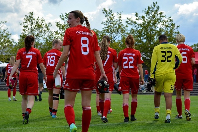 Sicht von hinten auf weibliche Fu&szlig;ballspieler in roten Trikots, die auf einem gr&uuml;nen Platz zum Spielbereich gehen.