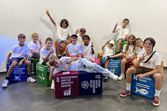 Gruppenfoto von Kindern in wei&szlig;en T-Shirts, die auf bunten Nachhaltigkeitsw&uuml;rfeln sitzen und freundlich posieren.