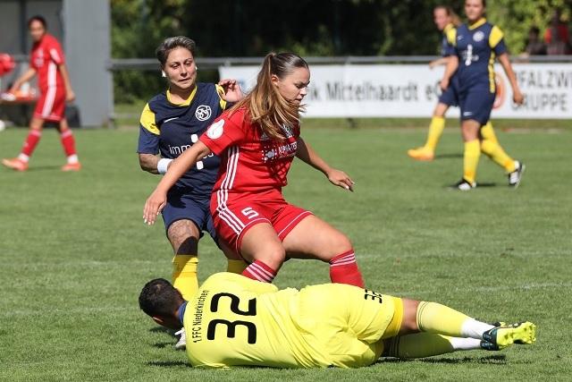 Spielszene im Fußball: Zwei Spielerinnen in roten und blauen Trikots kämpfen um den Ball, während ein Torwart am Boden liegt.