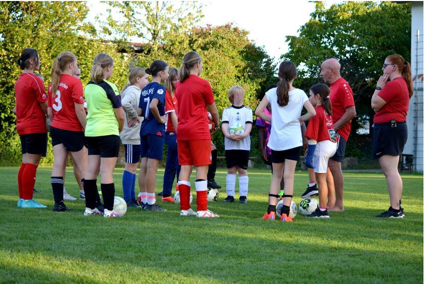 M&auml;dchenmannschaft versammelt sich um Trainer auf einem Fu&szlig;ballfeld, w&auml;hrend sie einer Anleitung zuh&ouml;ren.