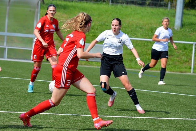Vier Frauen in Fu&szlig;balltrikots spielen auf einem Kunstrasenplatz, eine Spielerin dribbelt den Ball.