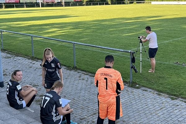 Frauenmannschaft in Trainingsanz&uuml;gen sitzt auf der Trib&uuml;ne, w&auml;hrend ein Fotograf das Fu&szlig;ballfeld aufnimmt.