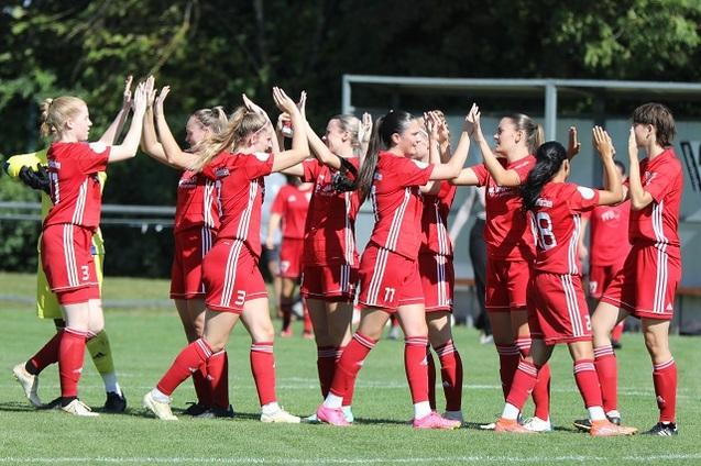 Gruppe von Spielerinnen in roten Trikots feiert auf einem Fu&szlig;ballfeld, umgeben von einer gr&uuml;nen Wiese.