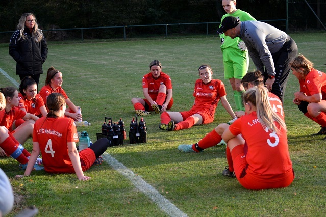 Frauenfu&szlig;ballmannschaft in roten Trikots sitzt im Kreis auf dem Rasen, w&auml;hrend der Trainer eine Taktikbesprechung h&auml;lt.