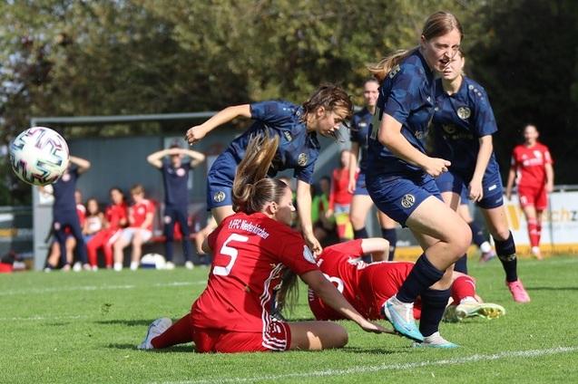 Zwei Fu&szlig;ballspielerinnen k&auml;mpfen um den Ball, w&auml;hrend eine weitere Spielerin auf dem Rasen liegt. Publikum im Hintergrund.