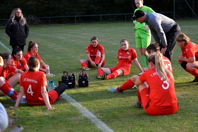 Frauenfu&szlig;ballmannschaft in roten Trikots sitzt im Kreis auf dem Rasen, w&auml;hrend der Trainer eine Taktikbesprechung h&auml;lt.