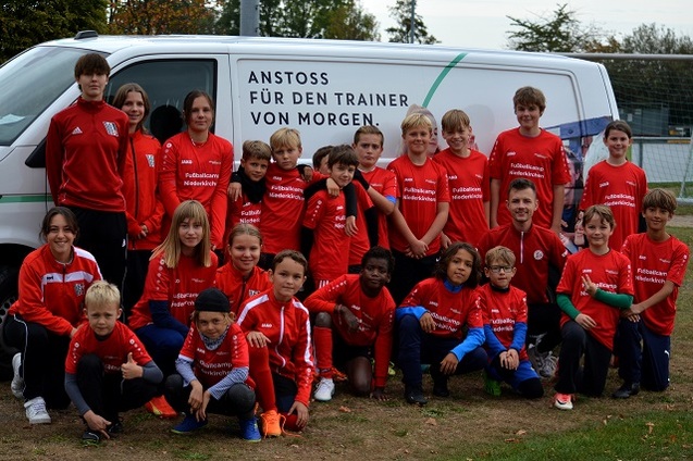 Gruppe von Kindern in roten Trainingsshirts steht vor einem Lieferwagen mit Werbung f&uuml;r einen Trainer-Workshop.