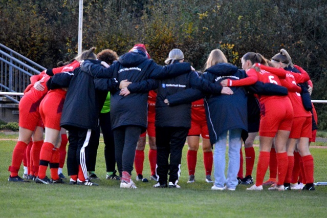 Gruppenumarmung eines Fu&szlig;ballteams in roten Trikots vor dem Spiel auf dem Rasenplatz.