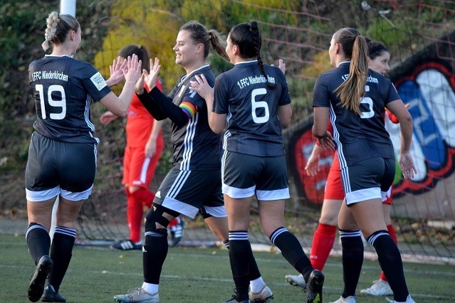 Vier Fu&szlig;ballspielerinnen in schwarz-wei&szlig; feiern den Torerfolg auf dem Feld, mit einem Spieler in rot im Hintergrund.