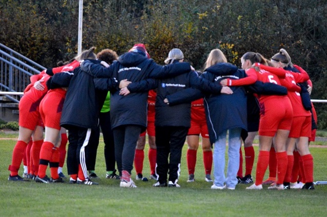 Gruppenumarmung eines Fu&szlig;ballteams in roten Trikots vor dem Spiel auf dem Rasenplatz.