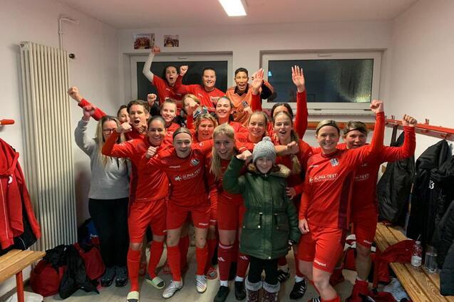 Gruppe von Fu&szlig;ballspielerinnen in roten Trikots posiert freudig in Umkleidekabine mit erhobenen H&auml;nden.