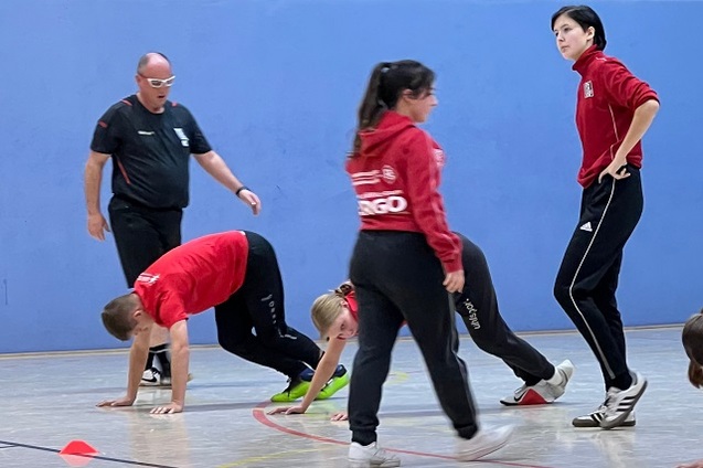 Gruppentraining mit Sportlern in roter Kleidung; ein Trainer beobachtet; Teilnehmer f&uuml;hren &Uuml;bungen auf dem Hallenboden aus.