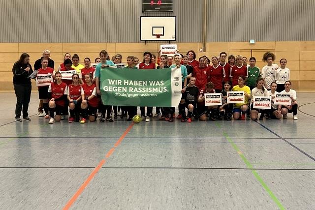 Gruppenshot von Fußballspielerinnen in roten und weißen Trikots, gemeinsam mit einem Plakat gegen Rassismus.