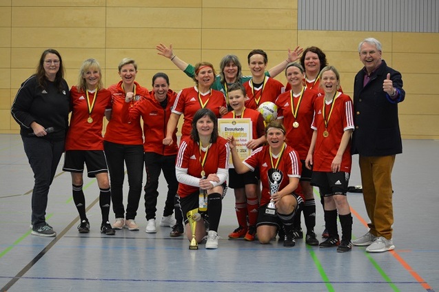 Gruppe von Frauen in roten Fu&szlig;balltrikots mit Medaillen, Pokalen und Urkunde, jubelnd in einer Sporthalle.