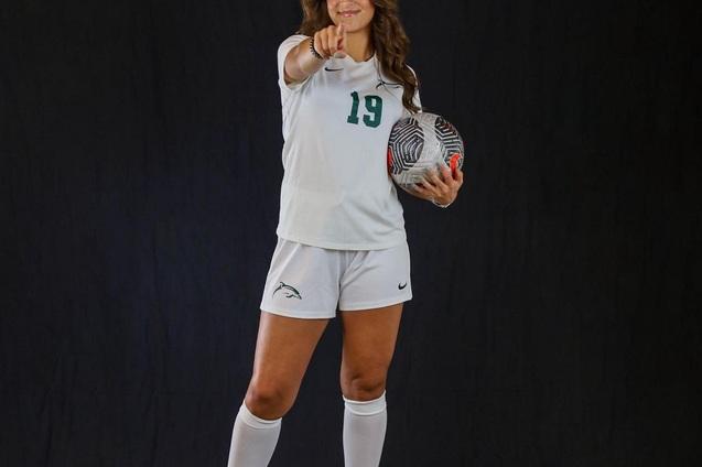 Junge Frau in Sportbekleidung h&auml;lt einen Fu&szlig;ball und zeigt mit dem Finger in die Kamera, vor schwarzem Hintergrund.