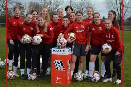 Gruppe von elf Fu&szlig;ballspielerinnen in roten Trainingsjacken mit B&auml;llen um einen runden Ballst&auml;nder auf einem Rasenplatz.