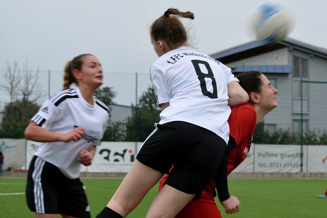 Zwei Fu&szlig;ballspielerinnen im Kampf um den Ball, eine springt &uuml;ber die andere in orangefarbener Torwartkleidung auf dem Spielfeld.