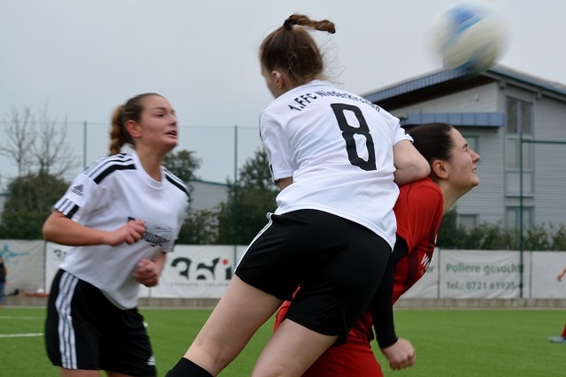 Zwei Fu&szlig;ballspielerinnen im Kampf um den Ball, eine springt &uuml;ber die andere in orangefarbener Torwartkleidung auf dem Spielfeld.