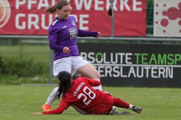 Zwei Fu&szlig;ballspielerinnen in Aktion, eine in lila mit gelocktem Haar, die andere in rot, im Zweikampf auf dem Rasen.