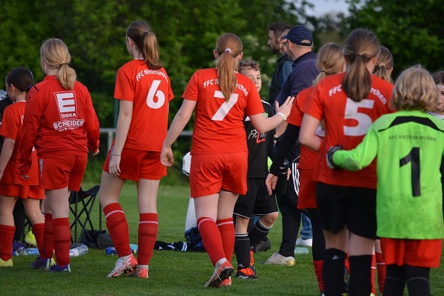 Gruppe von M&auml;dchenfu&szlig;ballerinnen in roten Trikots beim Verlassen des Spielfelds, Trainer beobachtet sie.