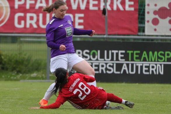 Zwei Fu&szlig;ballspielerinnen in Aktion, eine in lila mit gelocktem Haar, die andere in rot, im Zweikampf auf dem Rasen.