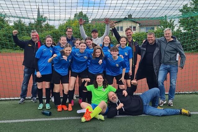 Gruppensfoto einer Fu&szlig;ballmannschaft mit Trainer, alle in blauen Trikots, vor einem Tor auf einem Sportplatz.