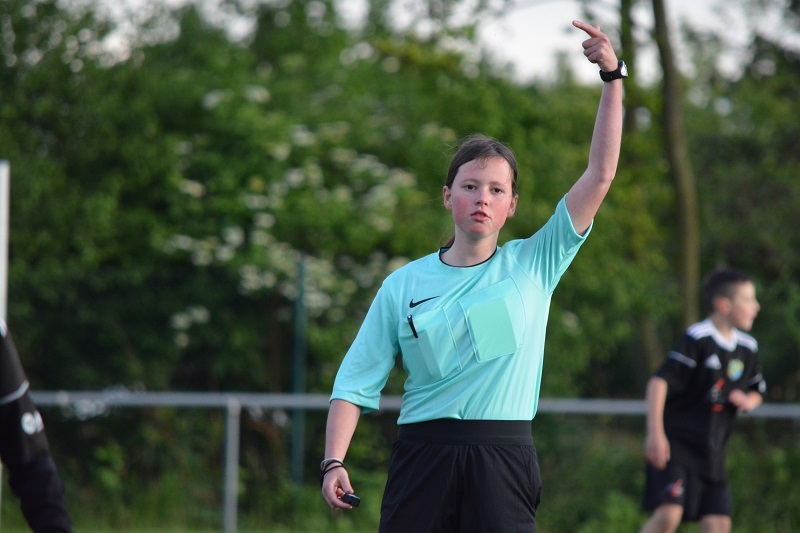 Schiedsrichterin in aquafarbenem Trikot zeigt mit dem Finger auf das Spielfeld w&auml;hrend eines Fu&szlig;ballspiels im Freien.