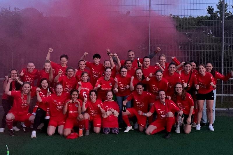 Gruppe feiernder Fu&szlig;ballspielerinnen in roten Trikots vor rosa Rauchfackeln auf einem Sportplatz.